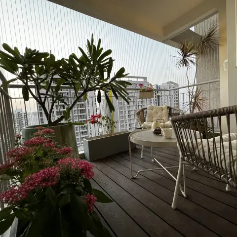 High-floor balcony decked in dark WPC boards, with a frangipani and pink ixora in grey planters against the invisible grilles and a rope-woven sofa and white