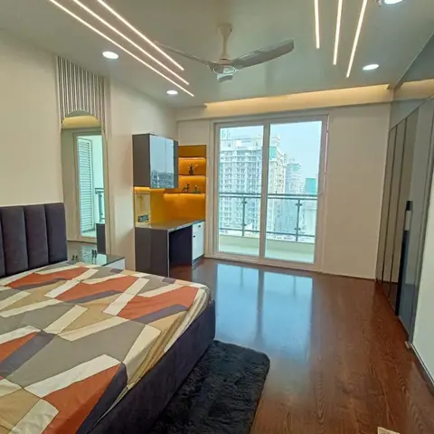 High-floor bedroom with a charcoal channel-tufted bed under a geometric orange quilt, facing a dark study-cum-dresser unit with a backlit mustard niche
