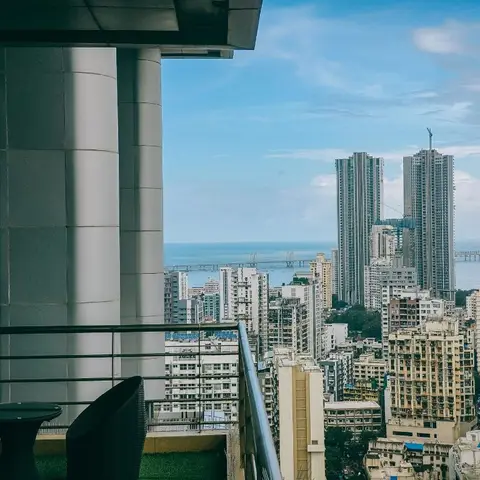 High-rise balcony with a round table and woven chairs overlooking a dense city and sea-link skyline.