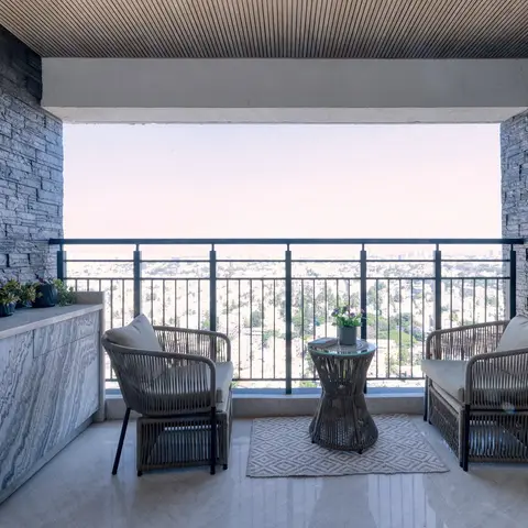 High-rise balcony with stacked-stone side walls, a linear wood-slat ceiling, two woven rattan lounge chairs around a round side table on a diamond-pattern rug