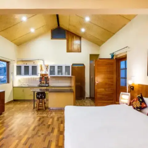 Hill-cottage studio under a woven bamboo pitched ceiling, a floral-cushioned bed at one end and a pistachio-green kitchenette with a bar stool at the other.