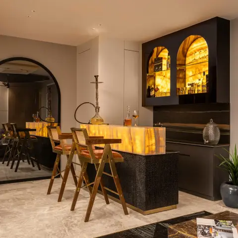 Home bar with a backlit honey-onyx counter on a fluted black base with brass toe-kick, twin arched onyx-lit liquor niches