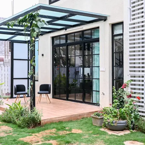 Home deck patio under a glass-roofed steel pergola with climbing vines, two black chairs, and black-framed glass sliding doors opening into the living room