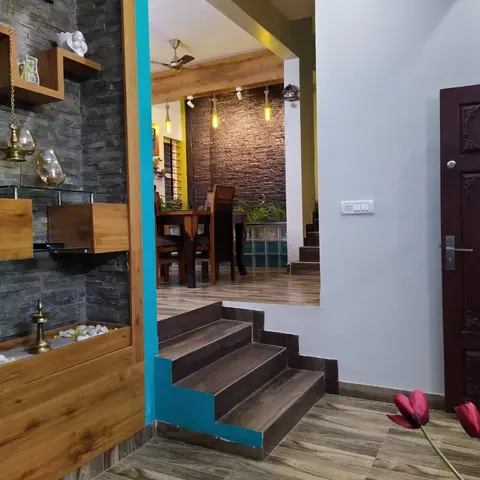 Home entrance foyer with a grey stone-clad wall, a wooden pooja display shelf, and a teal-accented step down into the dining area.