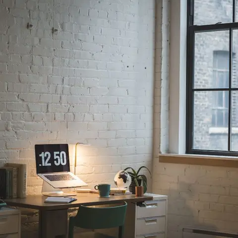 Home office nook against a whitewashed exposed brick wall with a wood desk, teal chair and a vintage typewriter beside a factory-style window.