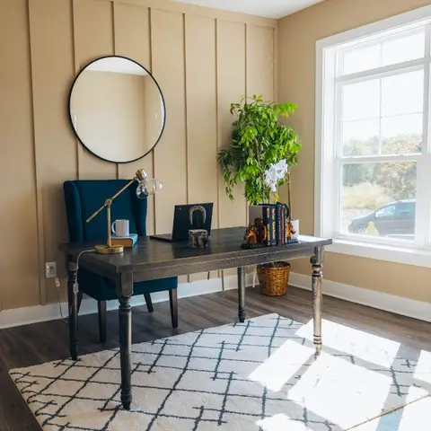 Home office nook with a board-and-batten tan accent wall, round mirror, teal wingback chair at a wood desk, and a large sunlit window.