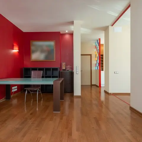 Home office nook with a deep red accent wall lit by sconces, a glass-top desk on a dark wood base, a black bookshelf/cabinet unit, and a framed painting