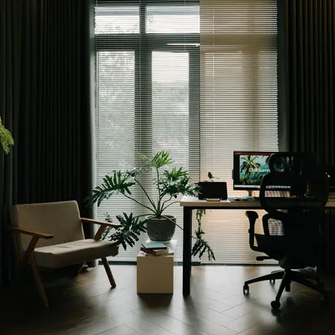 Home office nook with a mid-century wood-armed lounge chair, a dark wood desk with monitor, and floor-to-ceiling blinds behind.