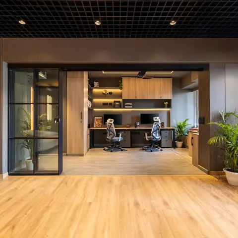 Home office nook with a two-seat wood desk setup for dual monitors, overhead wood cabinetry with backlit open shelving, a black grid ceiling