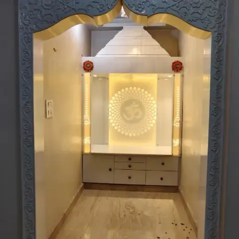 Home pooja room framed by a carved stone-effect arch with gold trim, a backlit Om-motif mandir unit with storage drawers below, marble flooring.