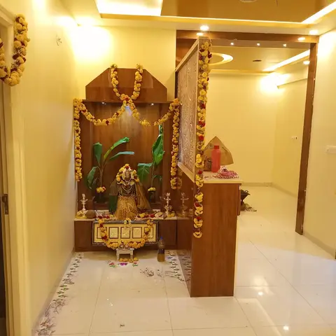 Home pooja unit with a carved wood mandir housing a garlanded deity idol, flanked by a jaali-carved wood partition, set in a foyer with cove-lit ceiling.