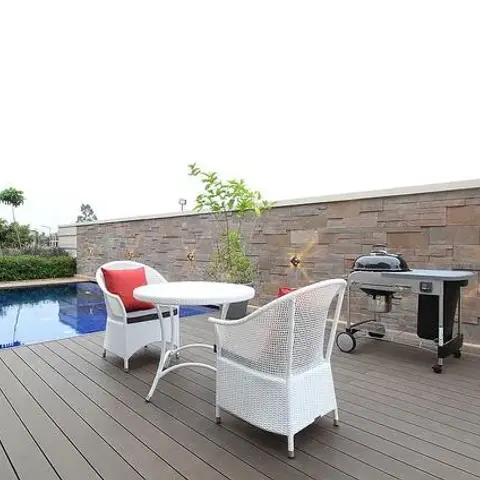 Home poolside deck with white wicker outdoor seating, red accent cushions, a gas grill, and a stacked stone privacy wall.