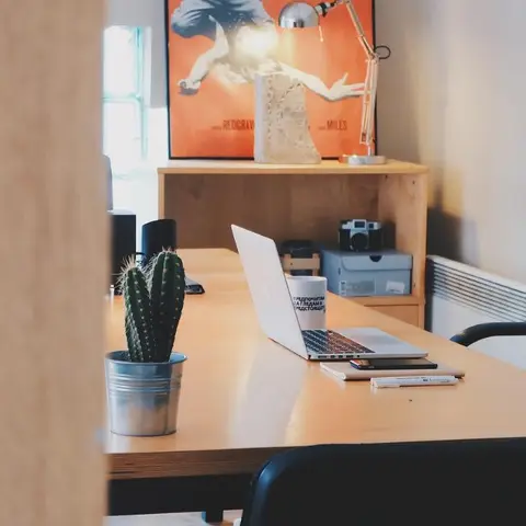 Home study desk with an open laptop, a small potted cactus and a framed vintage film poster on the shelf behind