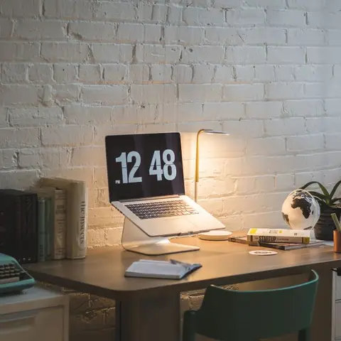 Home study nook against an exposed white-painted brick wall, with a wood desk holding a laptop on a stand, brass task lamp