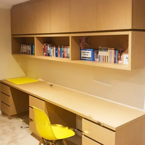 Home study nook with a full-width wood laminate desk, overhead cabinets and open book shelving, paired with a yellow molded chair.