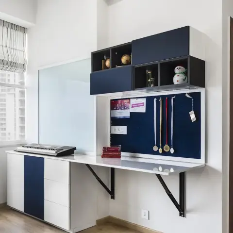 Home study nook with a wall-hung white desk leaf on black MS brackets, a navy pinboard hung with medals, and navy overhead cubbies beside a striped roman blind.