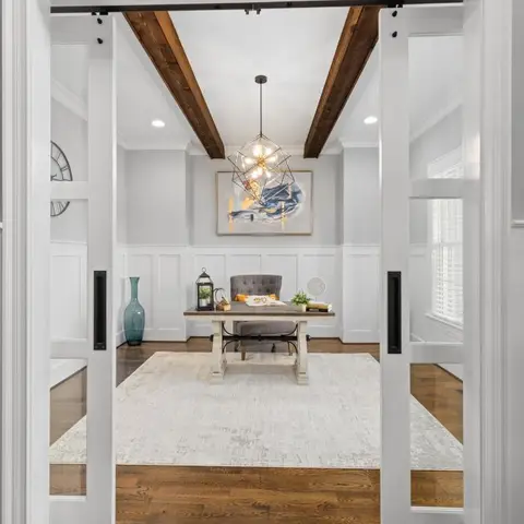 Home study seen through open glass barn doors, with a geometric wire pendant light, exposed wood ceiling beams, white wainscot panelling