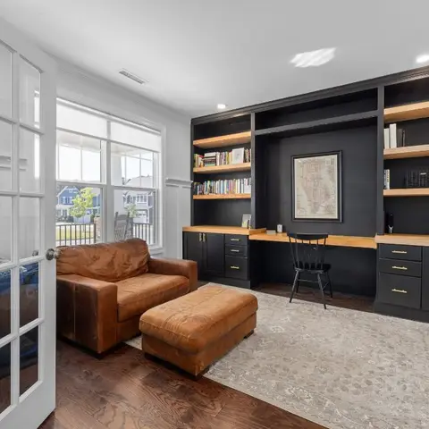 Home study with a black built-in bookshelf-and-desk wall trimmed in wood, beside a brown leather armchair and ottoman.