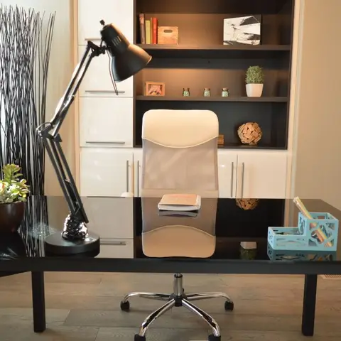Home study with a black glass-top desk, upholstered task chair, an adjustable desk lamp and a built-in two-tone bookshelf styled with books and decor.