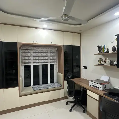 Home study with a long wood-topped desk under wall-hung black glass cabinets, facing a cream-and-black storage wall built around a cushioned window seat with a
