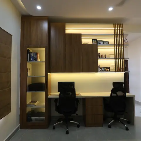 Home study with a two-seater built-in desk and overhead cabinets, a backlit open display shelf with framed photos, and a vertical wood slat screen on the right.