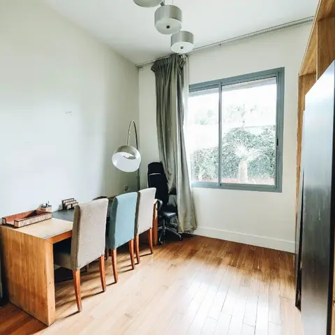 Home study with a wood desk and mismatched upholstered chairs, an arc floor lamp, sheer curtains, and a large abstract canvas leaning against the wall.