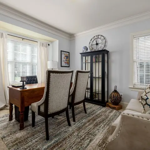 Home study with a wood writing desk, nailhead-trim upholstered chairs, a black curio cabinet and a patterned area rug under bay windows.