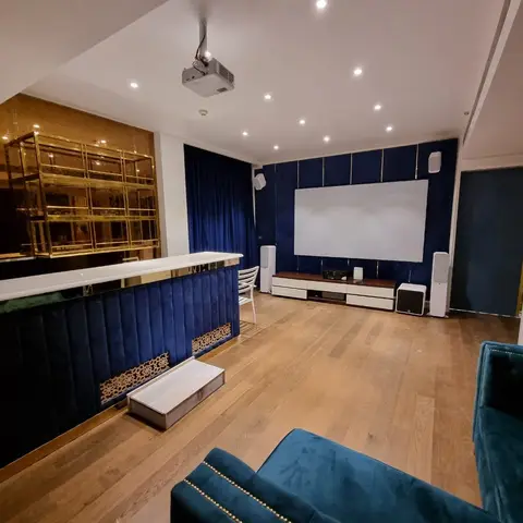 Home theatre and bar lounge with channel-tufted navy velvet wall panelling, a gold-framed glass bar shelf, a projector screen over a white media console