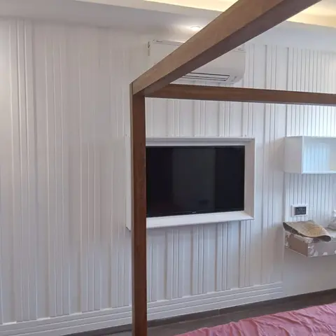Indian bedroom with a solid-wood four-poster canopy bed frame facing a wall-mounted TV set into fluted white wall panelling, cove-lit ceiling above.