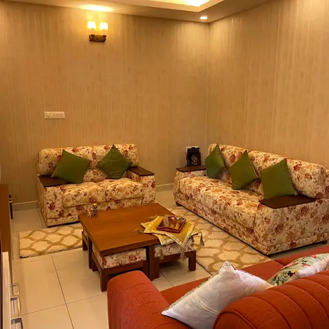 Indian living room with matching floral-print sofa sets, olive cushions, a wooden coffee table with nested stools, and a small pooja idol on a side table