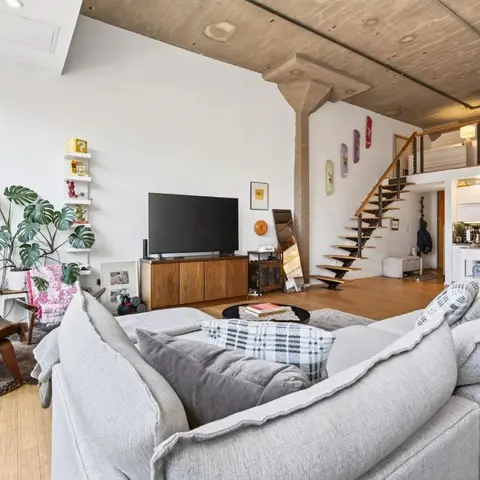 Industrial loft living room with an exposed concrete ceiling and column, a grey sectional sofa facing a wood media console, a spiral staircase to a mezzanine