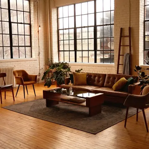 Industrial loft living room with exposed painted brick walls, tall steel-frame windows, a brown leather tufted sectional, mid-century wooden accent chairs
