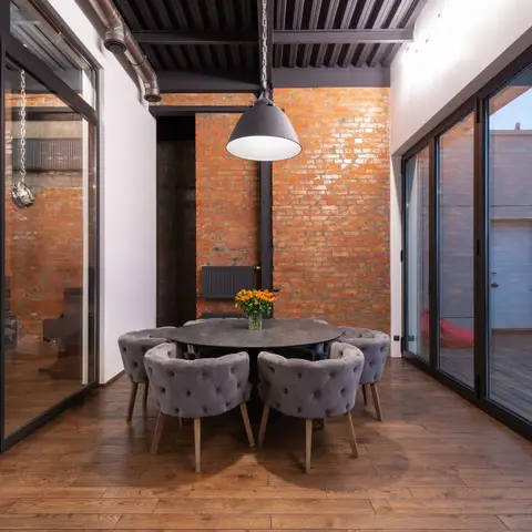 Industrial-style dining nook with exposed red brick wall, black exposed ceiling ducting and a large black dome pendant over a round table with tufted grey