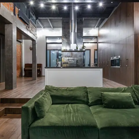Industrial-style open-plan living and kitchen with a dark emerald velvet sectional sofa in the foreground, a white kitchen island with steel chimney hood