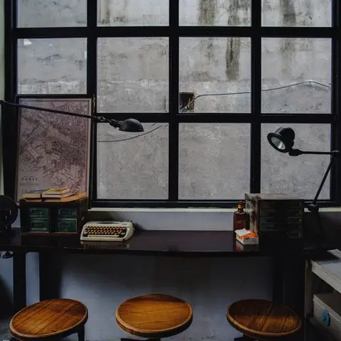 Industrial-styled study nook with a vintage typewriter and swing-arm task lamps on a dark wood desk facing a steel-frame window.
