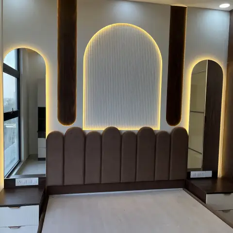 Just-handed-over bedroom, bed base still bare: a brown suede scalloped headboard runs wall to wall under a backlit fluted arch