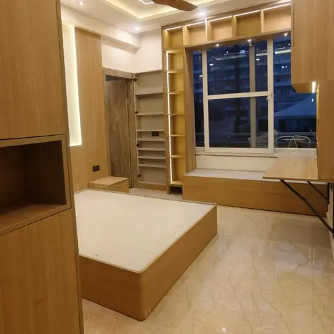 Just-handed-over bedroom in light-oak veneer at dusk, with a bare storage bed platform waiting for its mattress