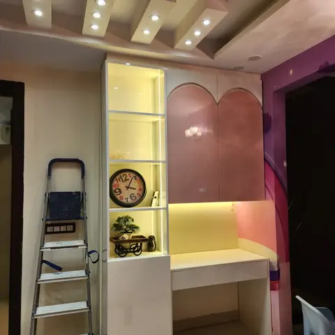 Kids' bedroom mid-installation with an arched pink-lacquered wardrobe, backlit display shelving, and a built-in study desk against a rainbow mural wall