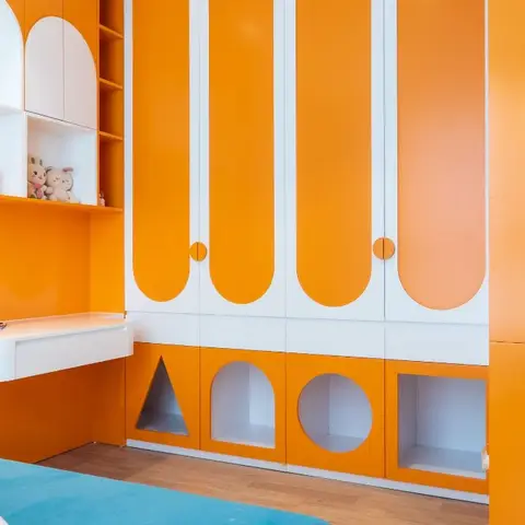 Kids' bedroom with a bright orange and white curved-door wardrobe wall featuring cutout play nooks, plus a built-in study desk and open toy shelving.