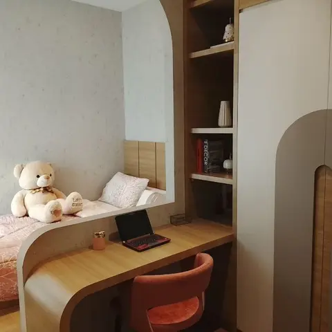 Kids' bedroom with a built-in curved study desk flowing into a low platform bed with pink bedding and a teddy bear, an arched wardrobe