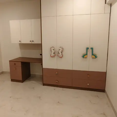 Kids bedroom with a peach-and-white wardrobe featuring unicorn and dinosaur decal appliques, plus a matching study desk and overhead cabinets.