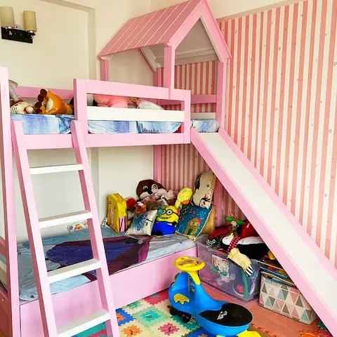 Kids bedroom with a pink playhouse-style bunk bed featuring a peaked roof canopy and slide, set against pink-and-white striped wallpaper