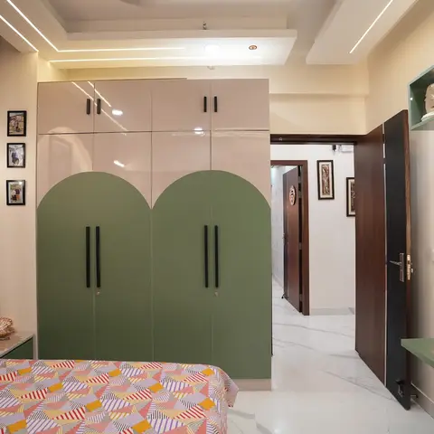 Kids' bedroom with an arch-cut two-tone cream and sage-green wardrobe, a geometric-pattern bedspread, and a TV playing a children's show through the open door.