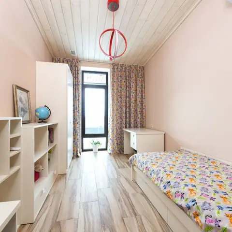 Kids' bedroom with pink walls, a cat-print bedspread, a white wardrobe and shelving unit, a red ring pendant light, and floral curtains by the balcony door.