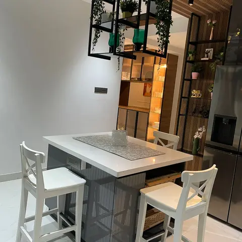 Kitchen island breakfast counter with three white cross-back bar stools beneath a hanging black-framed planter shelf