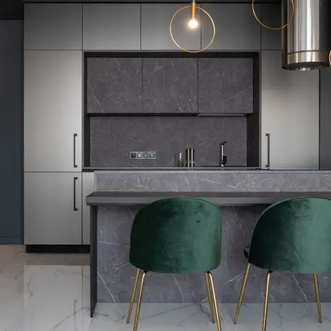 Kitchen island in book-matched grey marble with green velvet gold-leg bar stools, under a pair of circular gold-ring pendant lights.