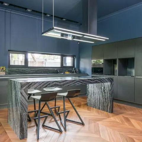 Kitchen island in dramatic dark waterfall-edge stone under a linear pendant hood, set against deep blue walls and herringbone parquet flooring.