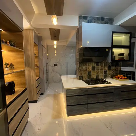 Kitchen island with a five-burner hob under a chimney hood, patterned tile backsplash, and a lit open wood shelving unit displaying decor along the passage.