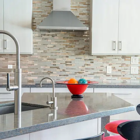 Kitchen island with a granite countertop, pull-down faucet, and a stainless steel chimney hood over a mosaic tile backsplash, with bar stools alongside.
