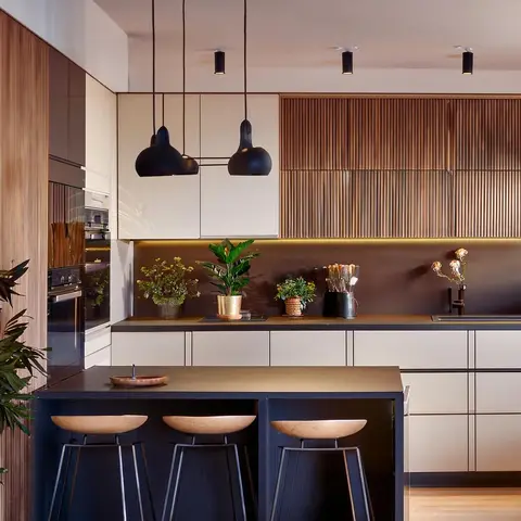 Kitchen island with black metal bar stools and three black dome pendant lights, facing a run of cream cabinetry and a fluted wood upper panel wall styled